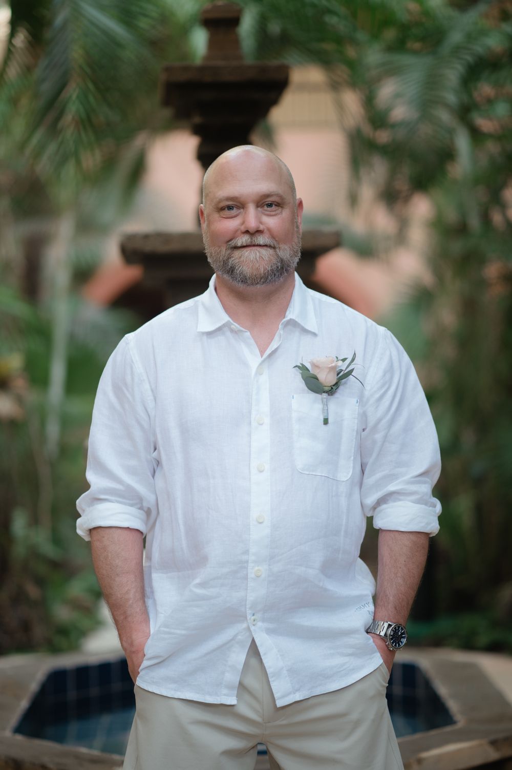 Person in white shirt and khaki shorts stands smiling in a tropical garden setting with stone fountain backdrop.