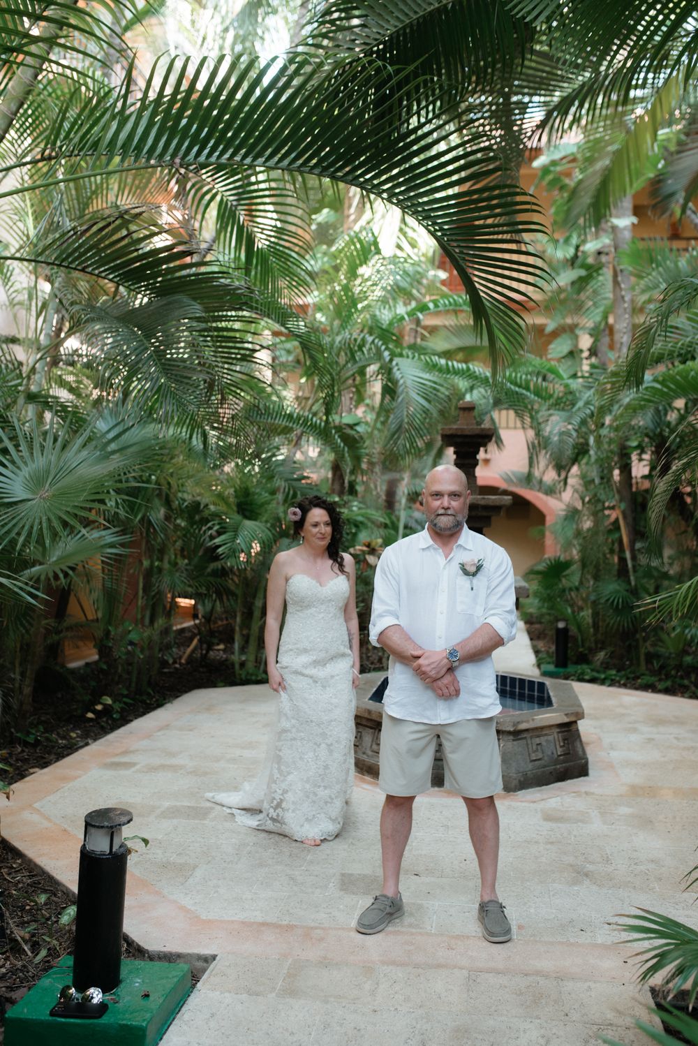 A wedding couple poses on a stone pathway surrounded by lush tropical palm trees and greenery at a resort.