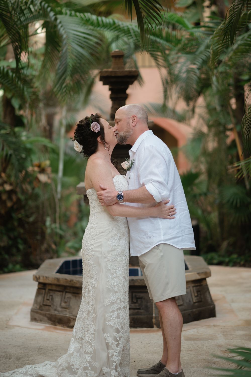 A romantic wedding couple share a kiss by a stone fountain surrounded by tropical palm fronds at an outdoor venue.