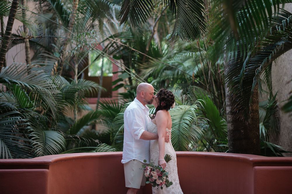 A romantic couple embraces on a terrace surrounded by lush tropical palm fronds and greenery.