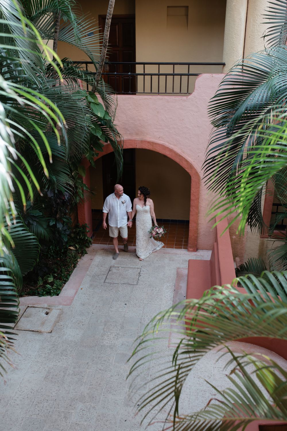 A couple walks along a tropical path with pink stucco walls and lush palm fronds framing the archway.