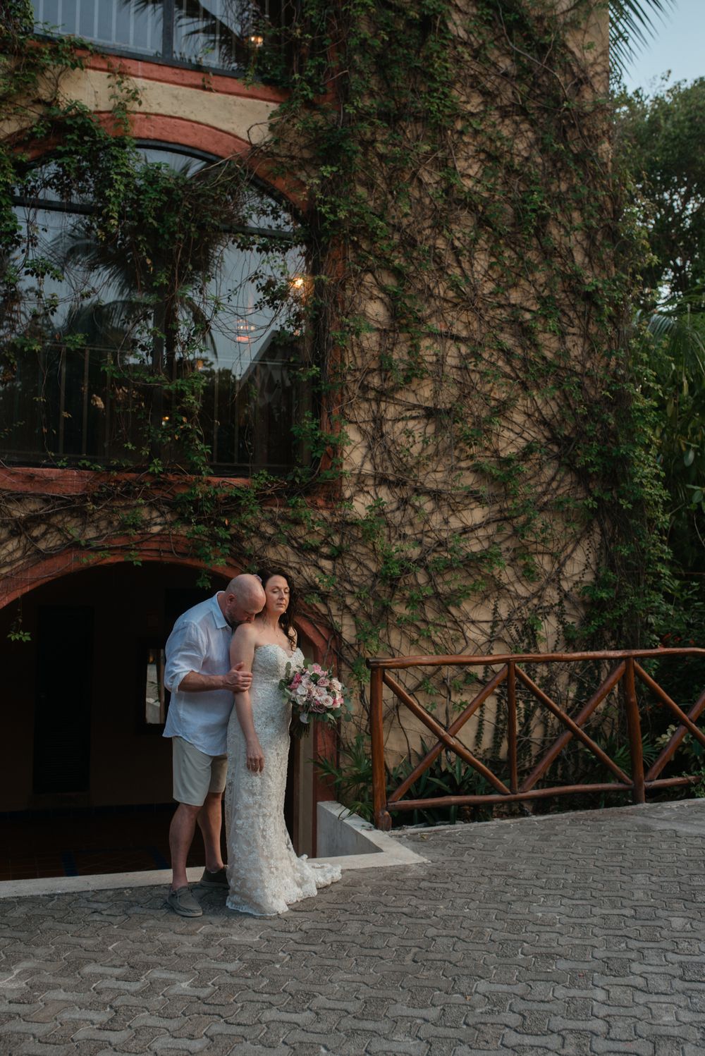 A romantic couple stands together at a stone building entrance with ivy covered walls and a wooden railing.
