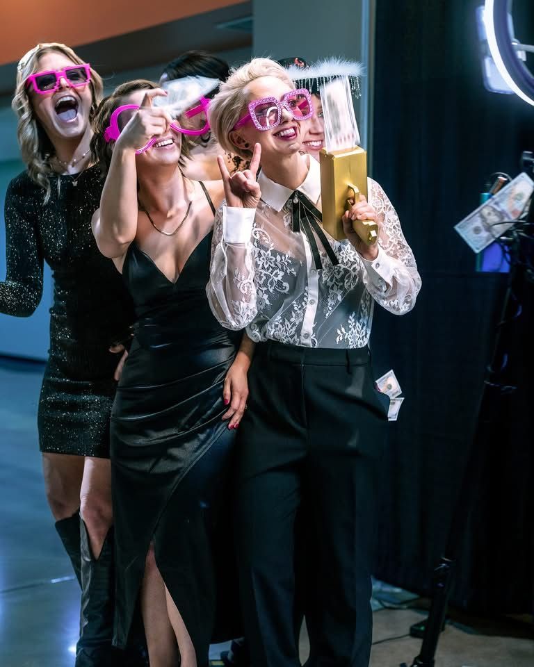 Three friends in pink sunglasses laugh and dance at a party while holding drinks and wearing festive outfits.