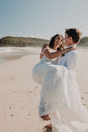 A joyful couple embraces and laughs together on a sunny beach with a long white dress flowing in the breeze.