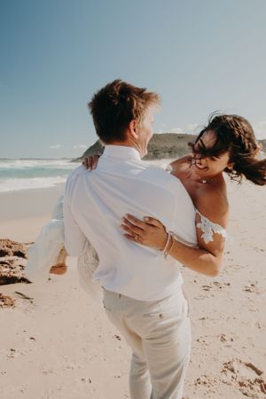 A couple shares a romantic moment while dancing together on a sunny beach with ocean waves in the background.