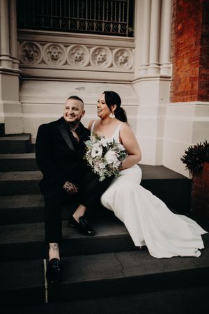 A wedding couple sits and laughs together on church steps, with the bride holding a white floral bouquet.