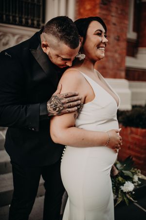 Candid wedding photo of couple embracing and laughing together outside brick building in classic formal attire.