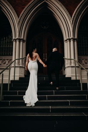 A bride in a sleek white backless gown ascends stone steps at an elegant Gothic church entrance with arched doorways.