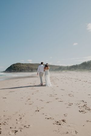 A couple walks together on a secluded beach with cliffs and blue sky in the background.