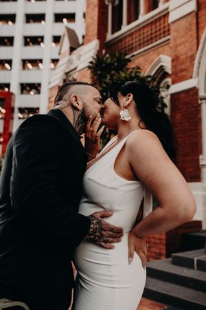 An intimate couple shares a romantic moment outside a brick building with modern architecture in the background.
