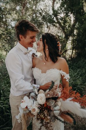 A romantic wedding couple embraces outdoors with a beautiful bouquet featuring white roses and dried flowers in natural sunlight.