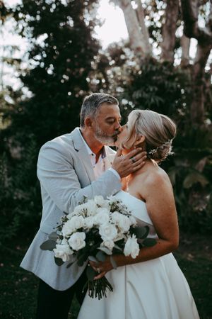 A romantic wedding moment captured outdoors with a beautiful white bouquet of flowers in a natural wooded setting.