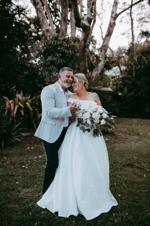 A couple shares a joyful moment outdoors at their wedding celebration, with the bride holding a white floral bouquet.