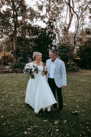 A couple shares a joyful moment in their wedding attire on a lawn surrounded by trees at dusk.