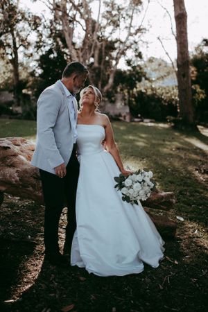 A wedding couple shares an intimate moment in a natural outdoor setting with trees and soft lighting at sunset.