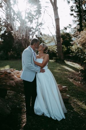 A couple in wedding attire share a romantic embrace in a sunlit wooded outdoor setting during their special day.