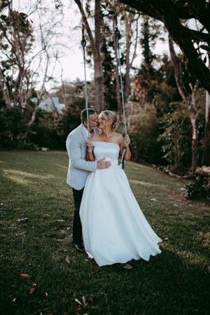 A couple in wedding attire embrace on a grassy lawn beneath tall trees at sunset casting a romantic silhouette.