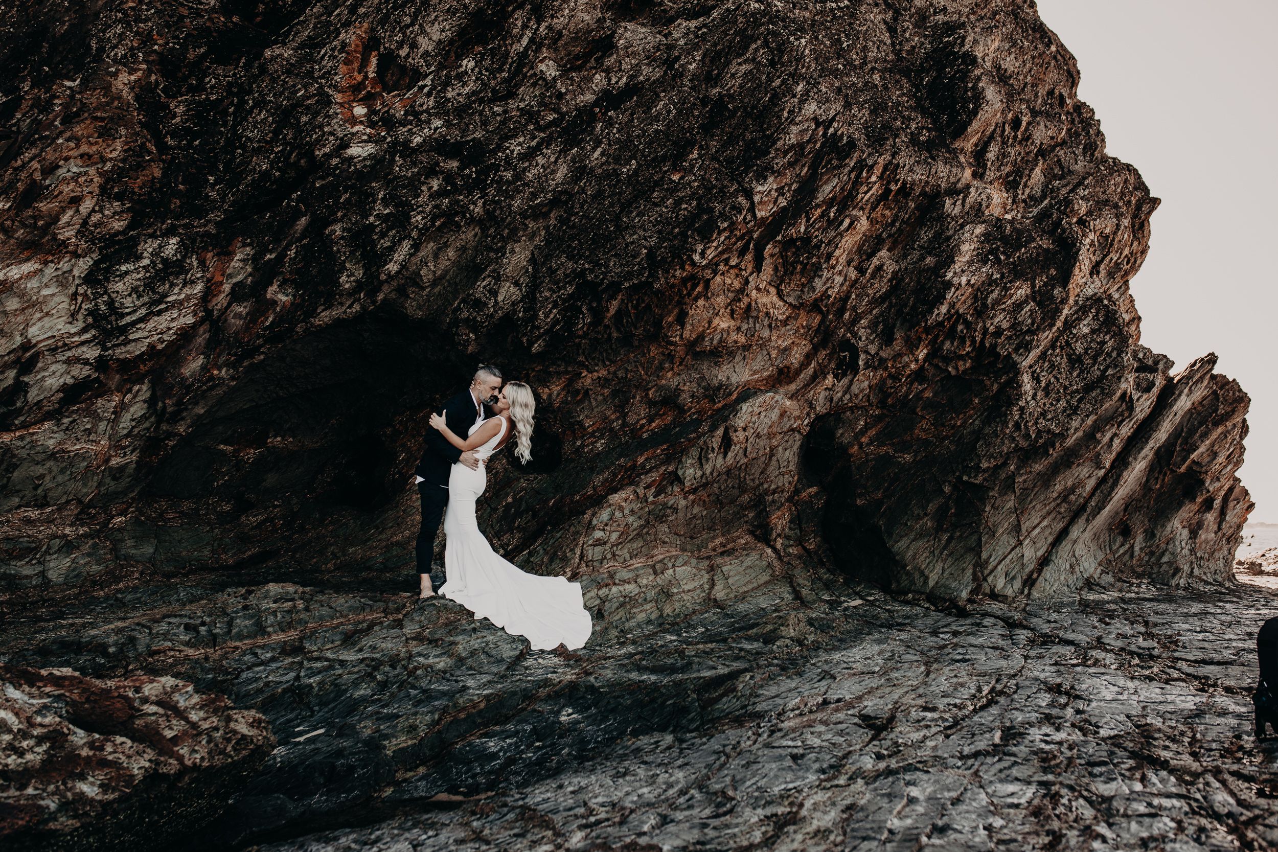 A person in a flowing white wedding dress stands dramatically against rugged rocky cliffs at sunset.