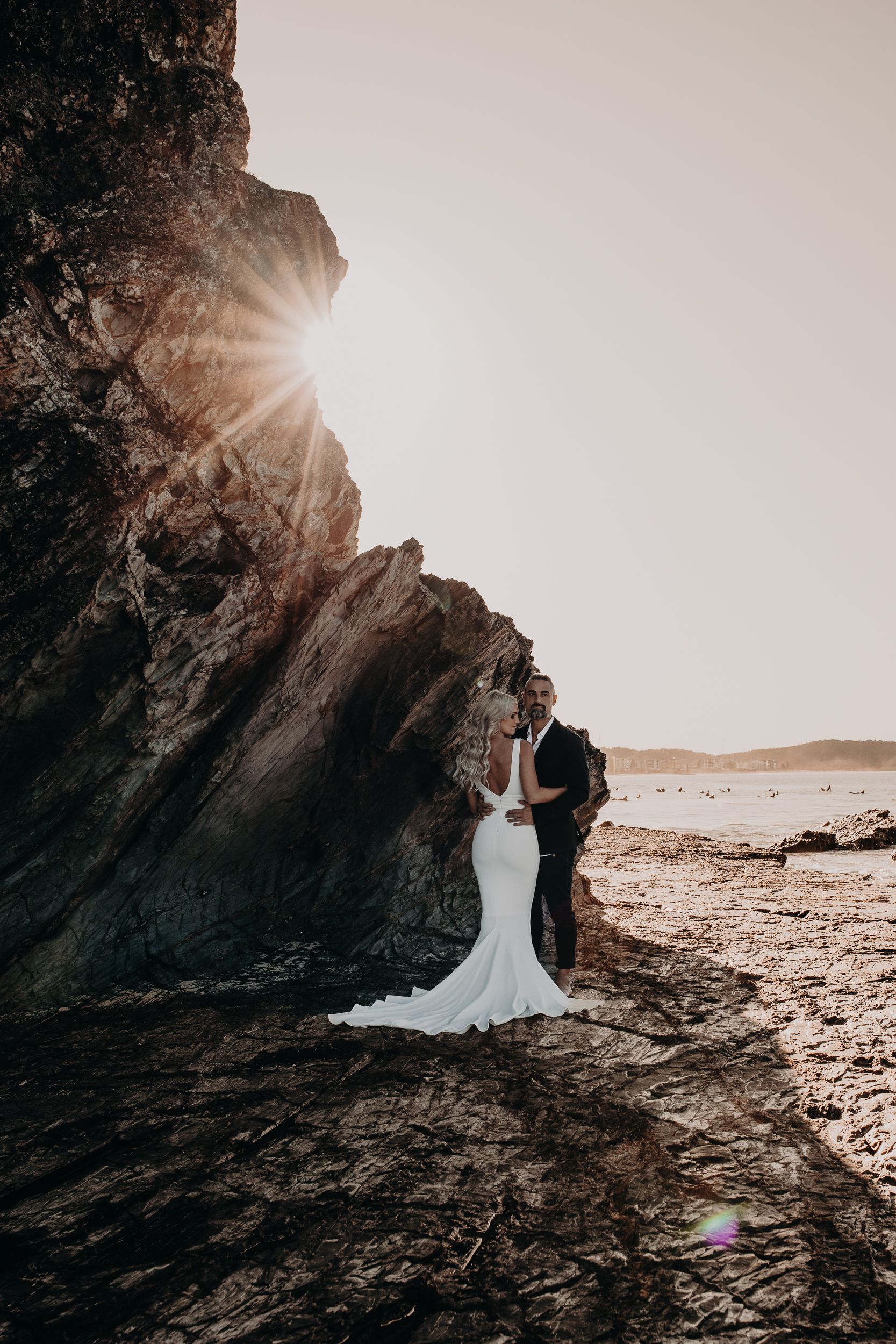 A couple embraces on a rocky beach at sunset with sunlight peeking through dramatic coastal cliffs.