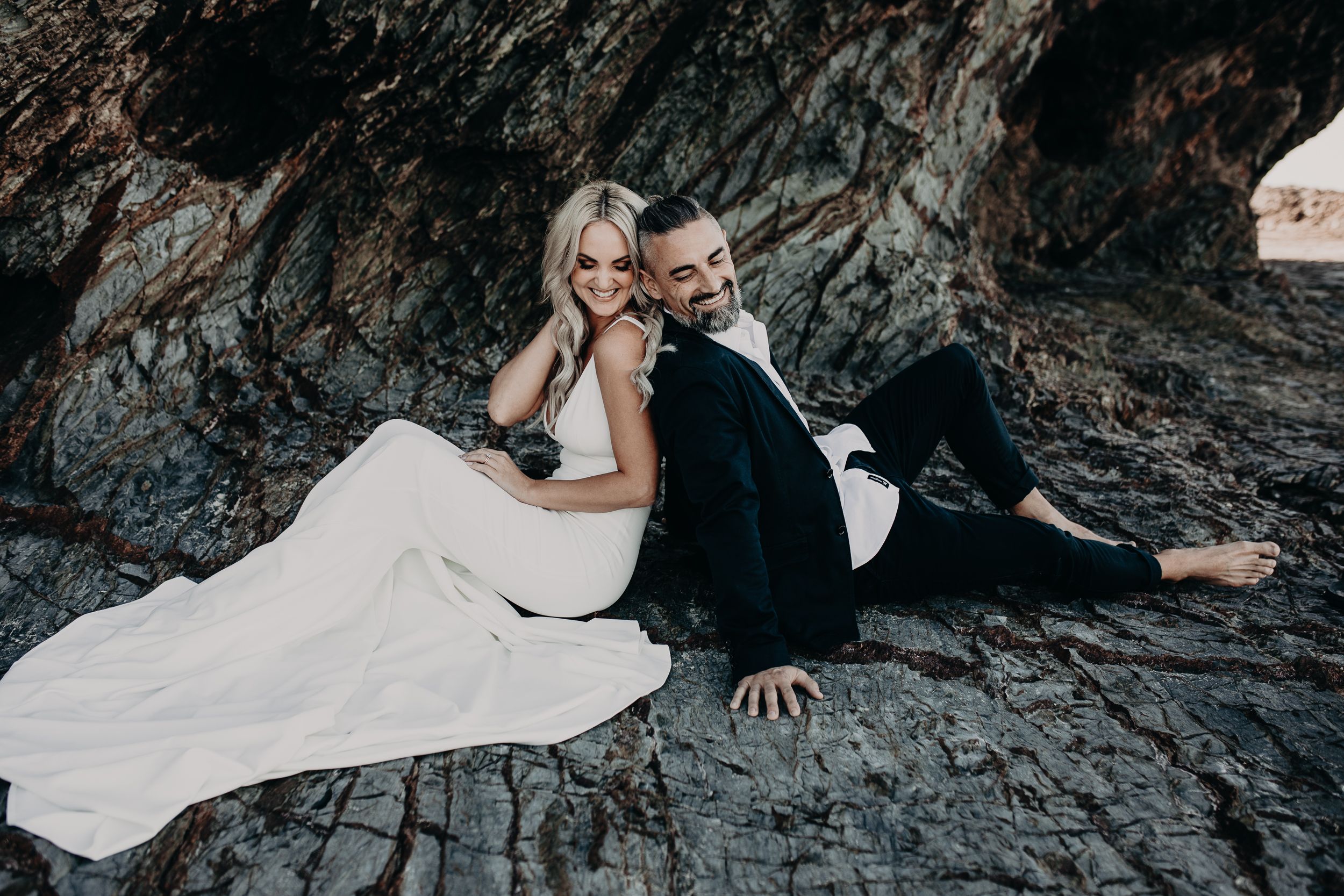 A couple in formal wedding attire reclines against dark rocks in a moody, atmospheric outdoor setting.
