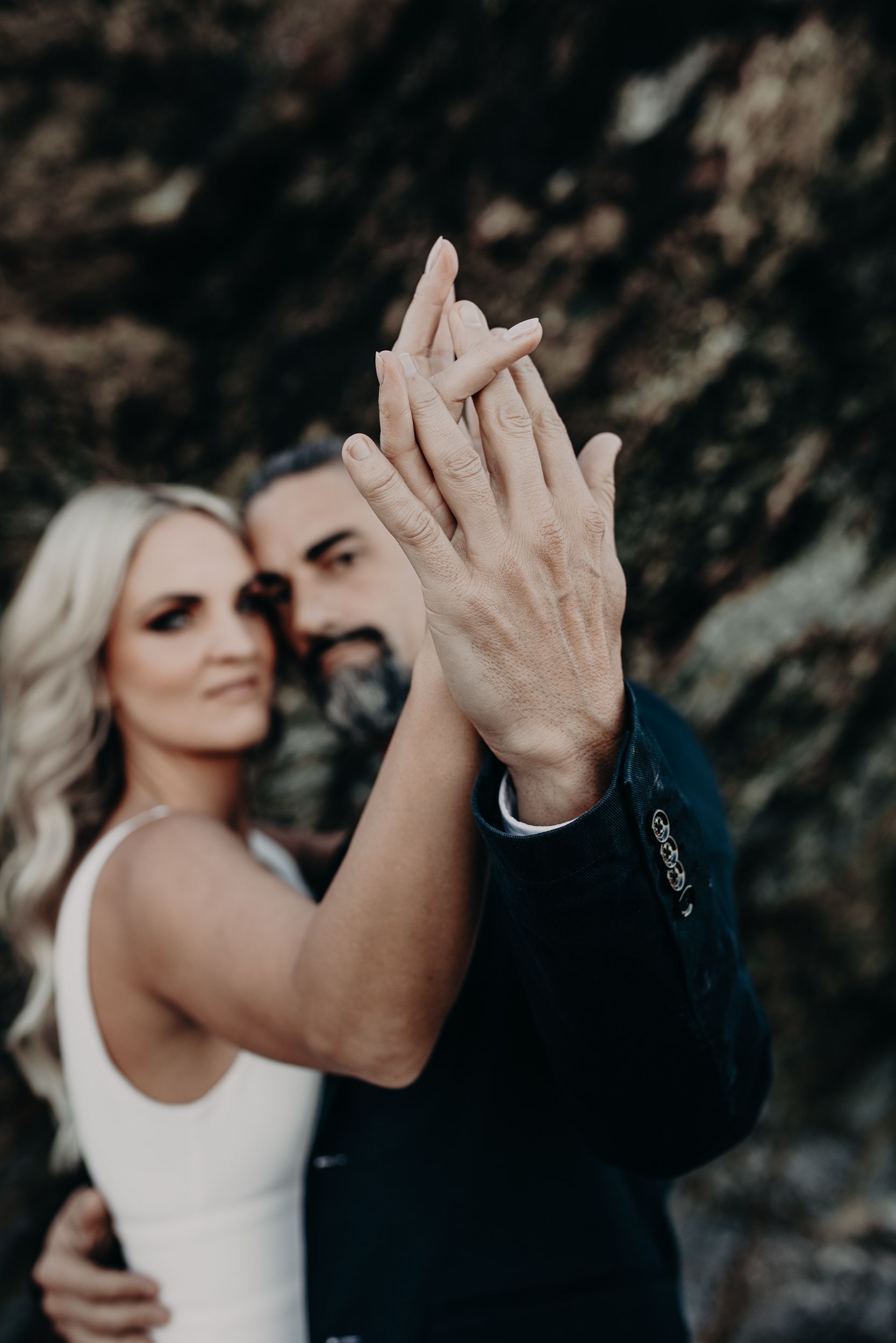 Close-up photo of a couple holding hands during their wedding engagement shoot against a dark rocky background.