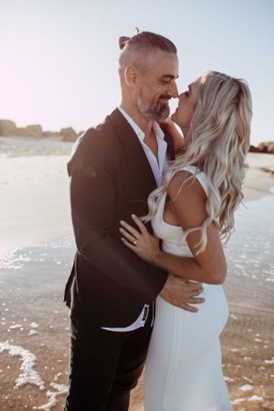 A romantic couple embraces on a sunlit beach, with the person in black holding the person in white close at sunset.