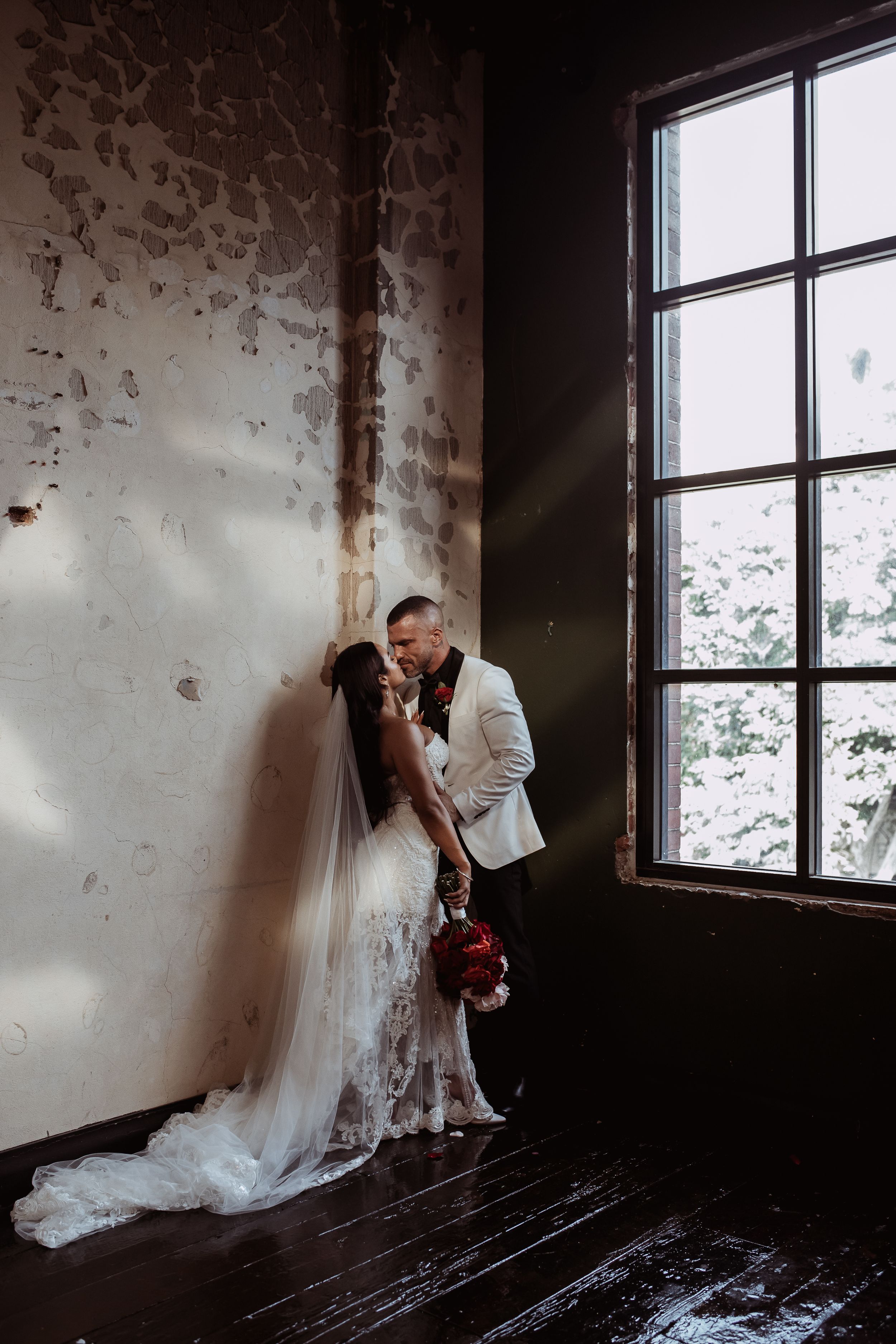 A romantic wedding couple shares an intimate moment by a large window in an industrial style room with peeling wallpaper.
