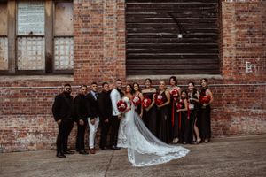Wedding party stands together against rustic brick wall backdrop with bride in flowing veil and burgundy bridesmaids dresses.