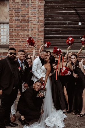 Wedding party in black attire poses with red bouquets against brick wall and wooden door at urban venue.