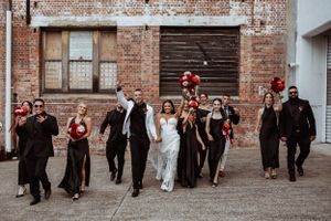 A wedding party dressed in black and white attire walks together in front of an industrial brick building exterior.