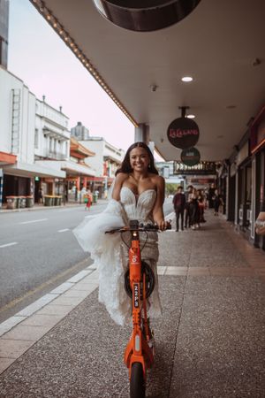 Person wearing a white wedding dress rides an orange rental bike on a city sidewalk under covered storefront walkway.