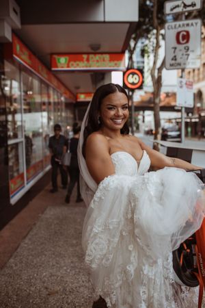 A bride in a beautiful white strapless lace wedding dress smiles while standing on a city street near storefronts.