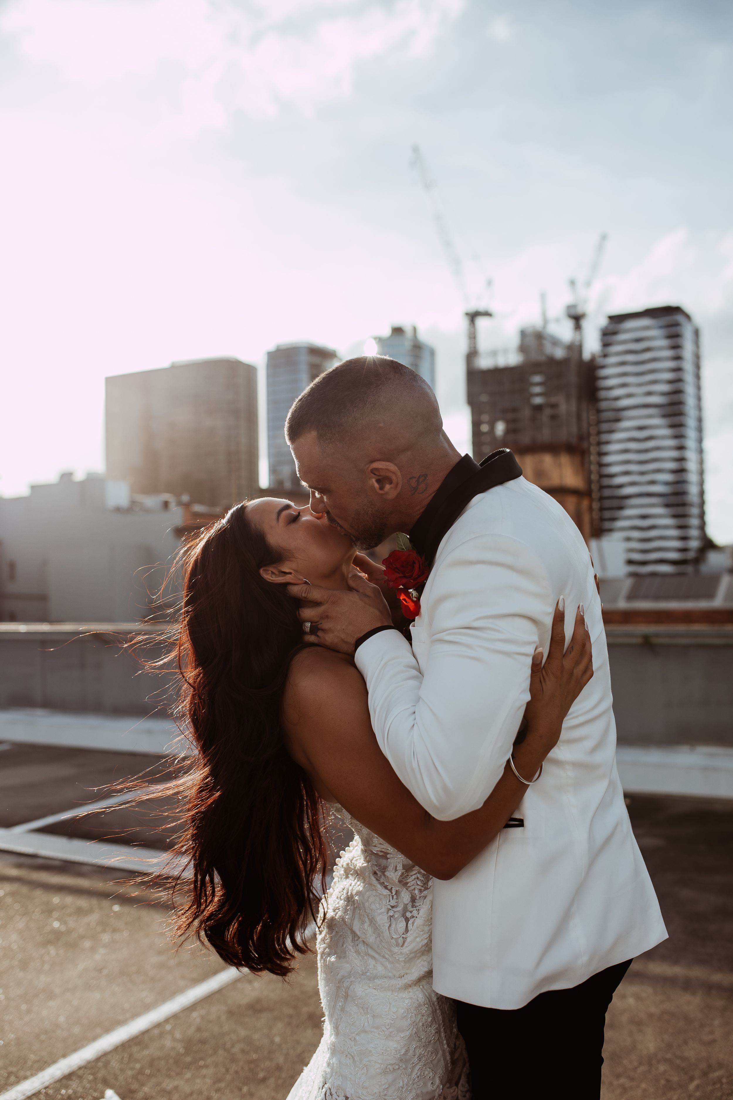 A romantic wedding couple share a passionate kiss on a rooftop with an urban cityscape backdrop at sunset.