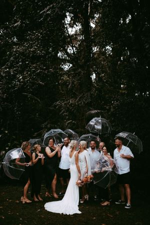 A wedding party holds clear umbrellas while gathered outdoors in a moody forest setting.