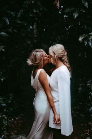 Two people in white dresses share an intimate moment in a lush garden setting.