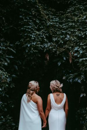 Two people in white wedding dresses hold hands while standing against a dark green leafy backdrop.