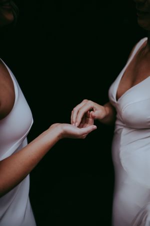 Two people in white dresses hold hands tenderly, shown in an intimate close-up shot against a dark background.