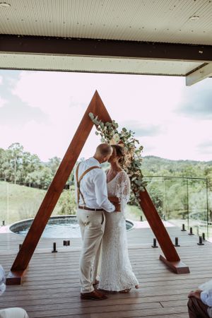 Wedding couple shares an intimate moment under a triangular wooden arch on an elevated deck overlooking scenic hills.