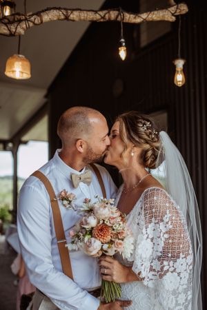 Romantic newlyweds share a tender kiss under rustic lighting while bride holds a delicate bouquet of blush and white flowers.