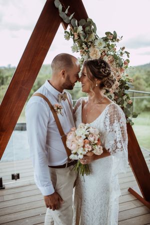 Romantic wedding couple stands beneath wooden arch decorated with eucalyptus and flowers on scenic outdoor deck.