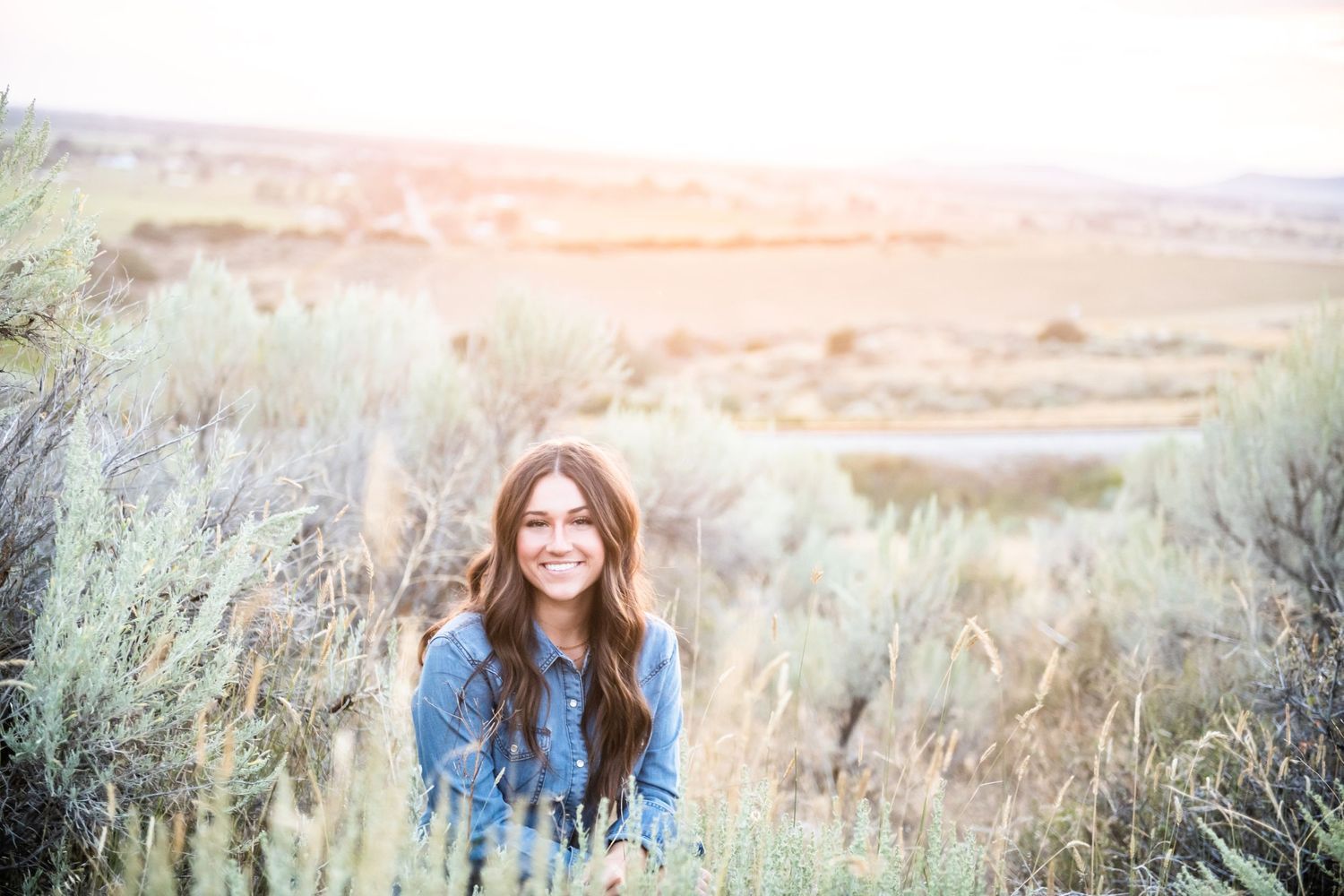 Person sits in tall grass with rolling hills and a sunset vista in the background.