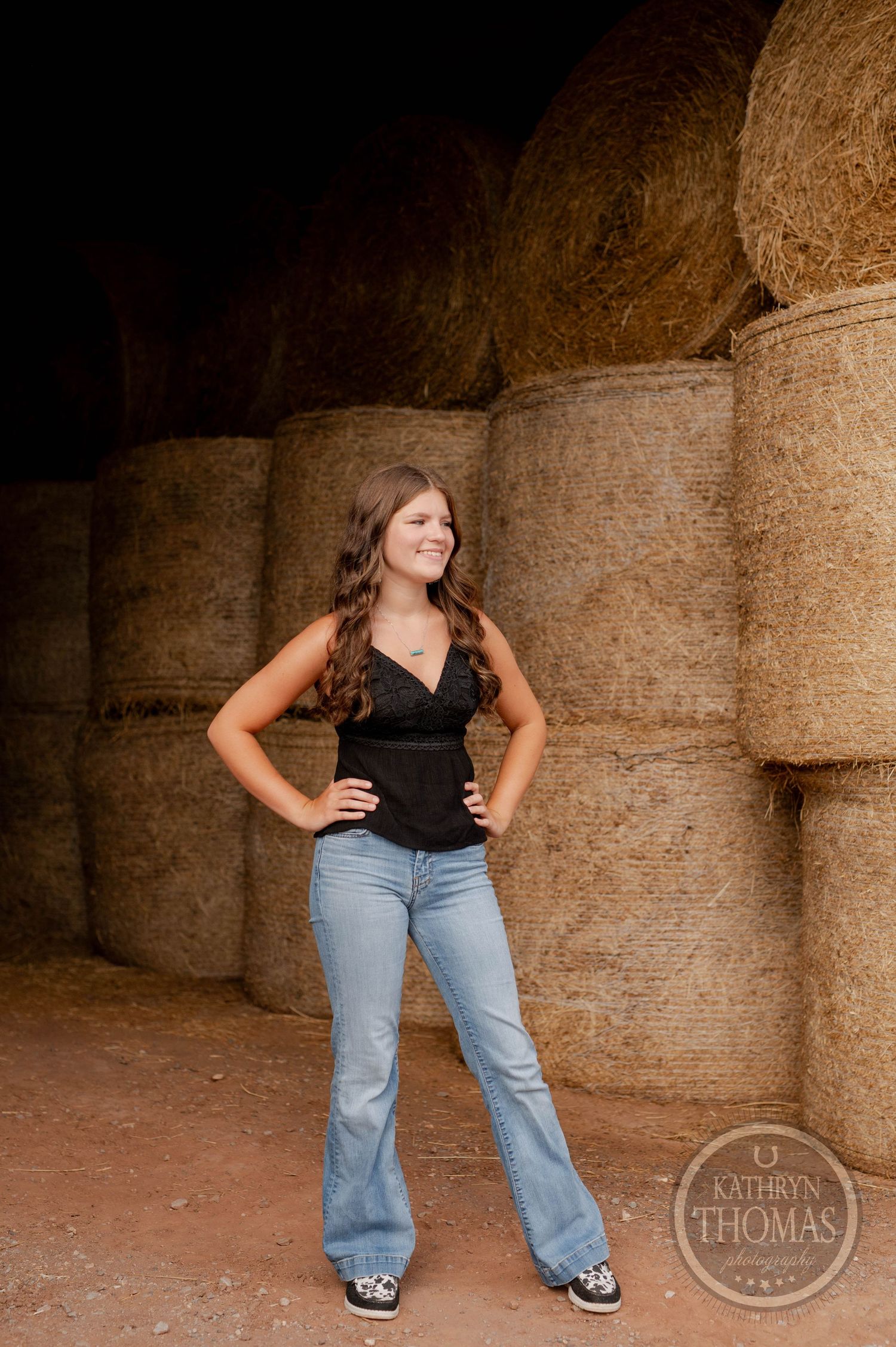 A person poses against hay bales wearing a black top and flare jeans in a rustic farm setting.