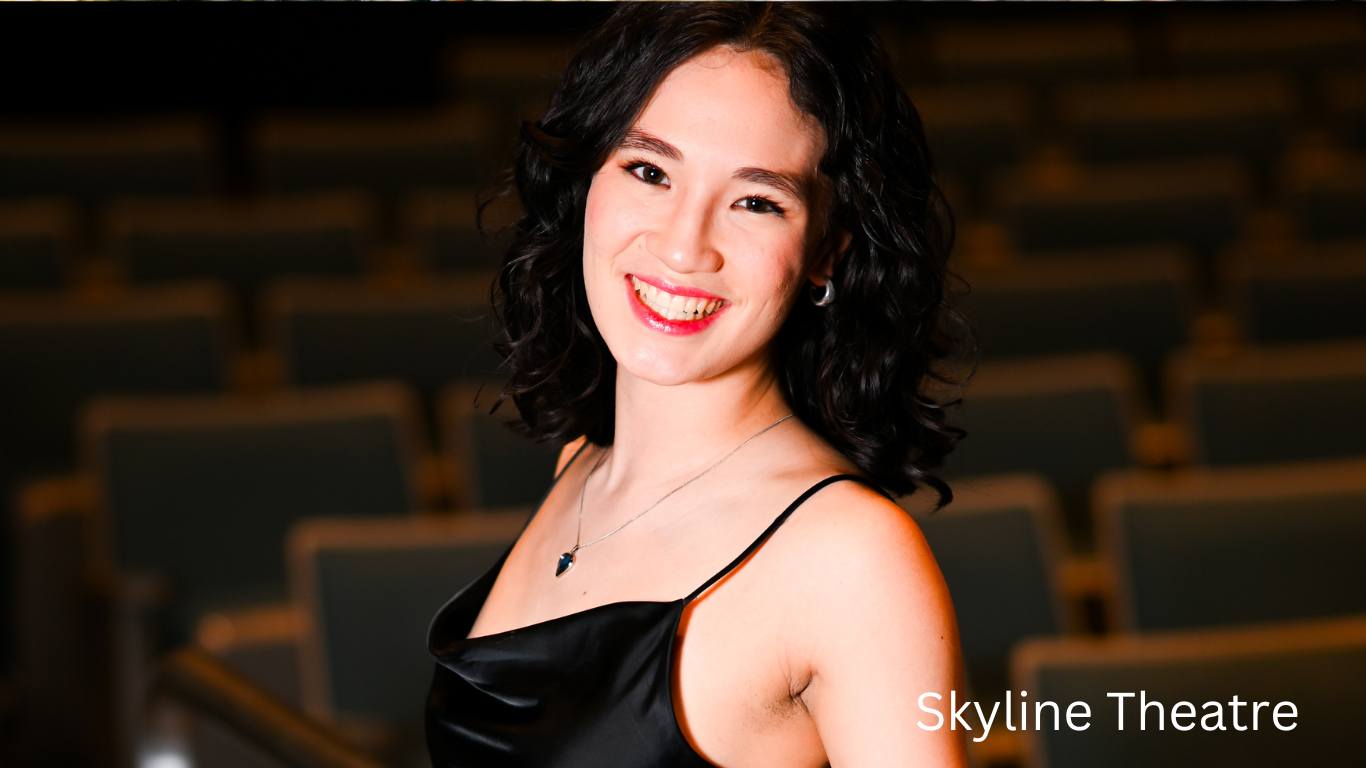 Performer in black evening dress poses elegantly against dark theatre backdrop.
