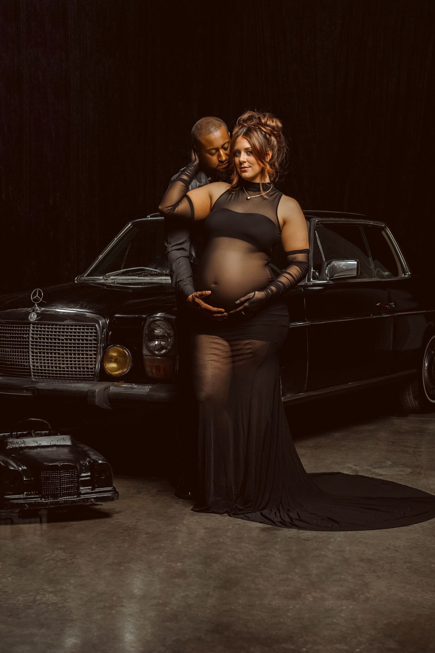 A couple poses by a classic black Mercedes-Benz for an elegant maternity photo shoot in a dark studio setting.