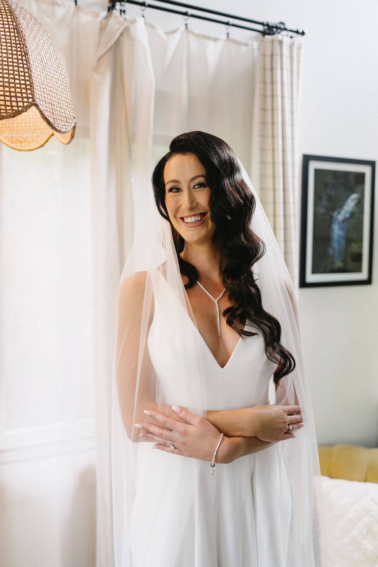 Beautiful bride in white v-neck wedding dress poses by window with flowing veil and long dark wavy hair.