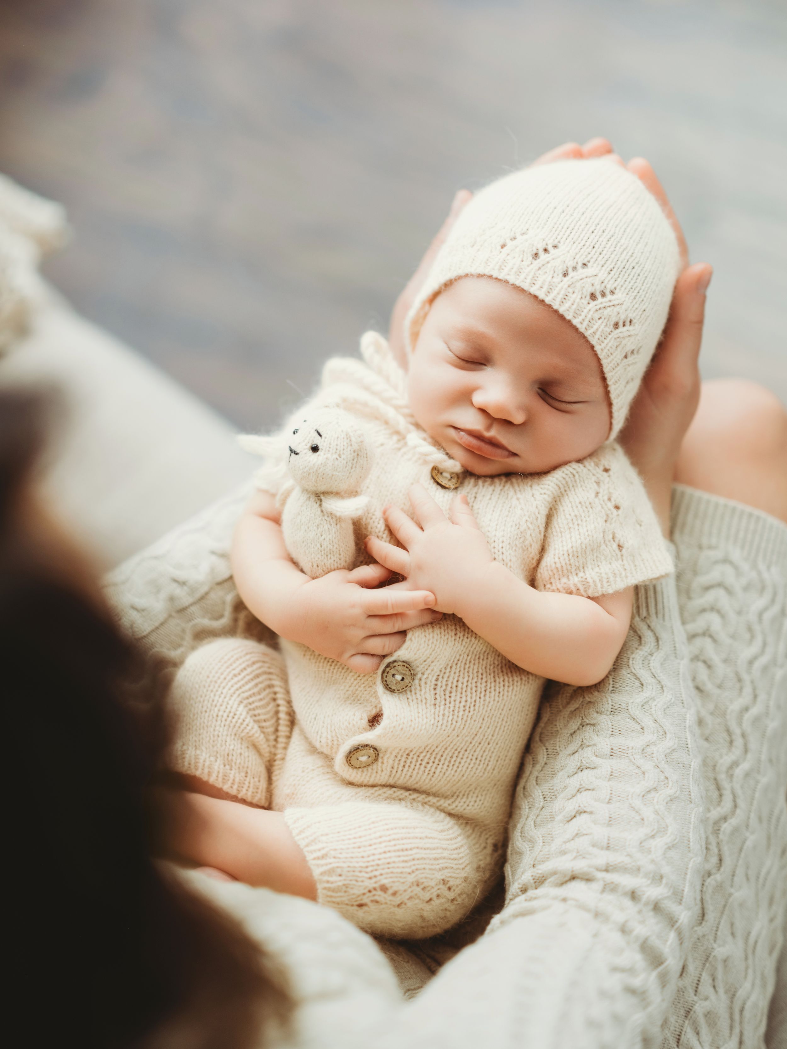 Sleeping newborn baby wearing cream knitted outfit and bonnet while holding small stuffed toy in cozy setting - Bellevue newborn photograph.