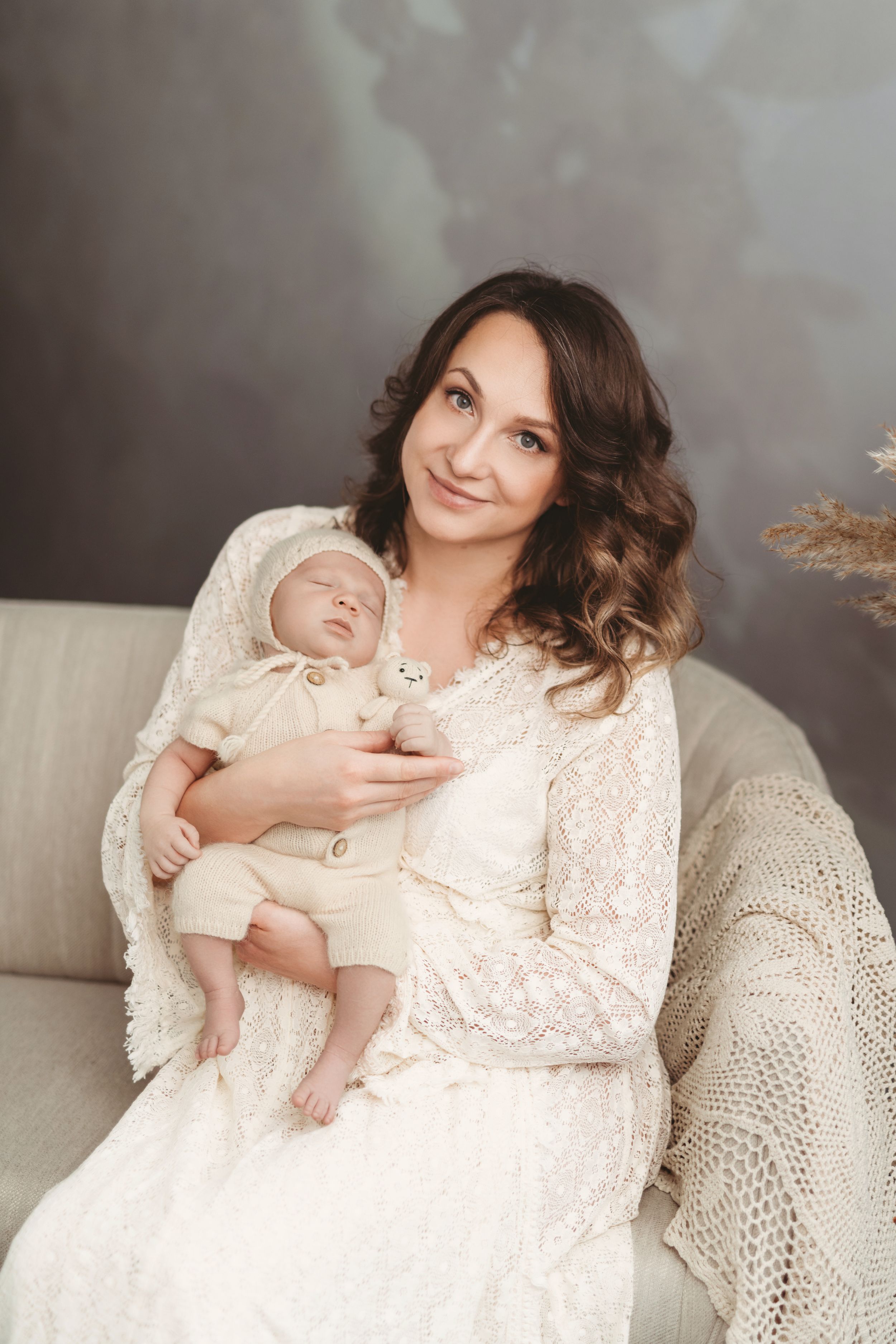 Mother in white lace dress holds sleeping newborn on couch with soft gray backdrop. | Bellevue, WA