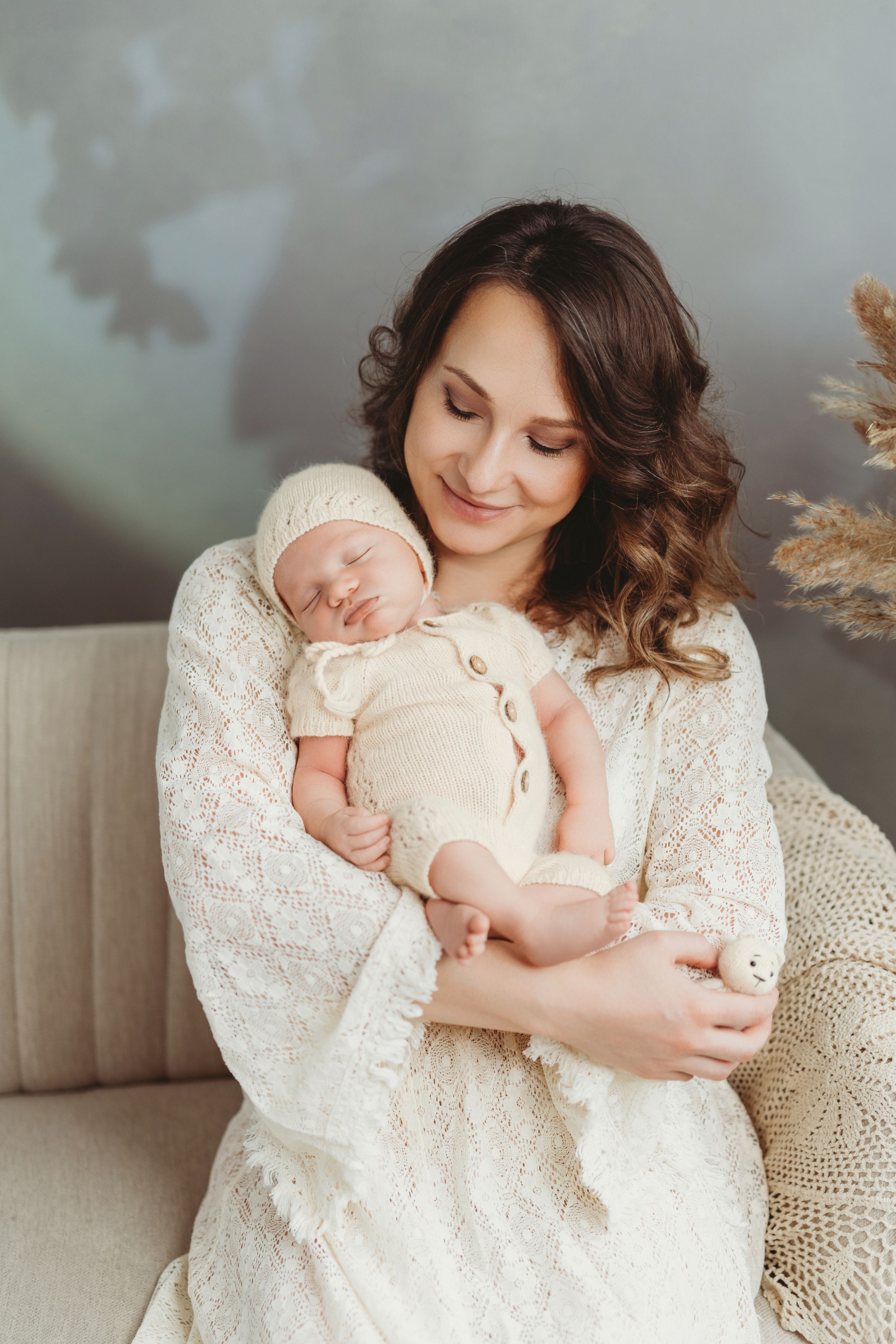 A tender moment captured as a woman in white lace holds a sleeping newborn on a beige couch with pampas grass decor. | Bellevue, WA