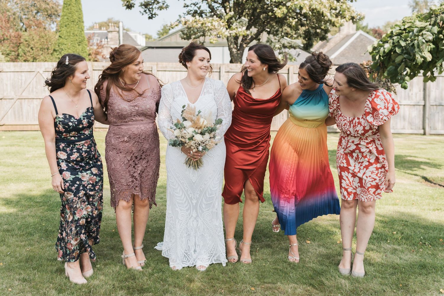Bridesmaids in colorful floral and solid dresses stand together on a sunny lawn during an outdoor wedding celebration.