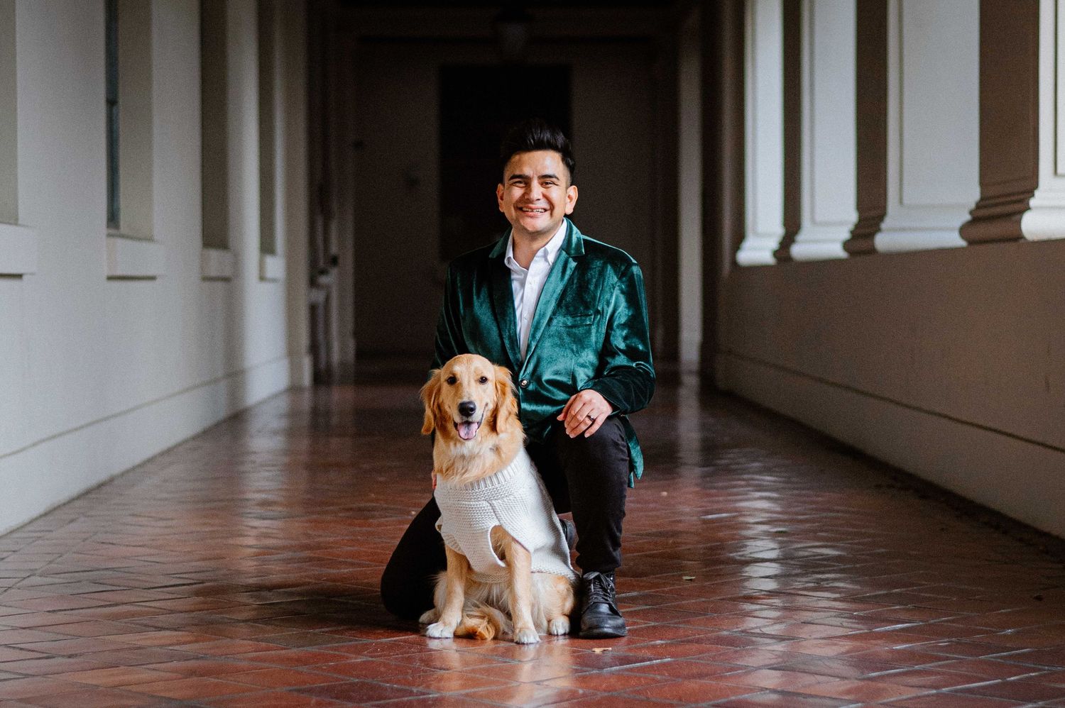 Person in green jacket poses with golden retriever in columned hallway.